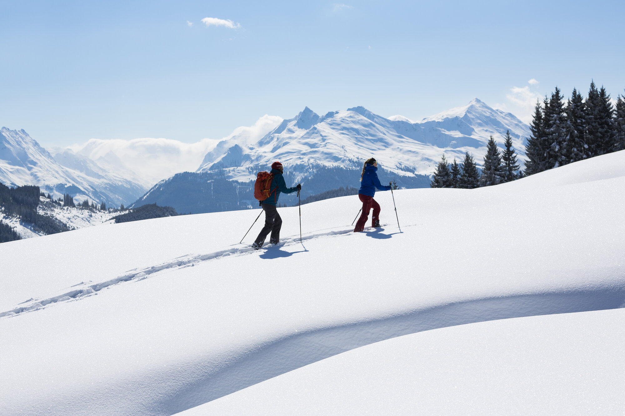 Eine Frau und ein Mann gehen hintereinander mit den Schneeschuhen einen verschneiten flachen Hang bergauf. Im Hintergrund sieht man tief verschneite Berge. Der Himmel ist blau und die Sonne scheint. 